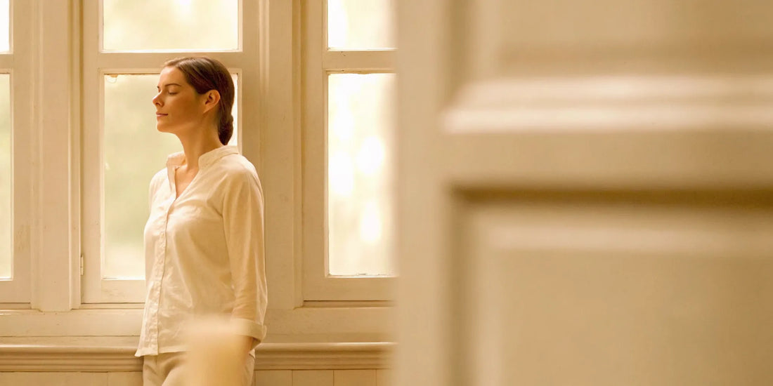 Woman standing by a window with soft natural light, calm and thoughtful expression representing mental clarity and wellness