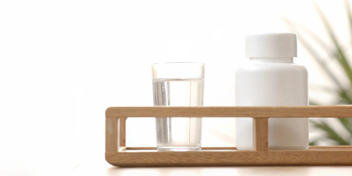 White postbiotic supplement bottle beside a glass of water in a sunlit modern wellness setting