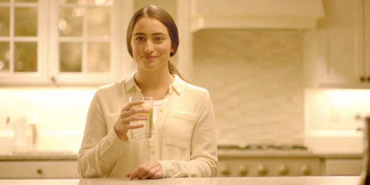 Calm woman in her 30s standing in a bright kitchen, holding a glass of water, representing digestive wellness and bloating relief