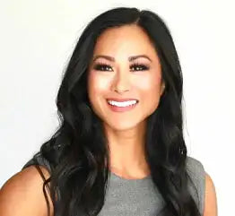 A woman with long, wavy black hair and a bright smile is wearing a sleeveless gray top. She is posing in front of a plain white background.