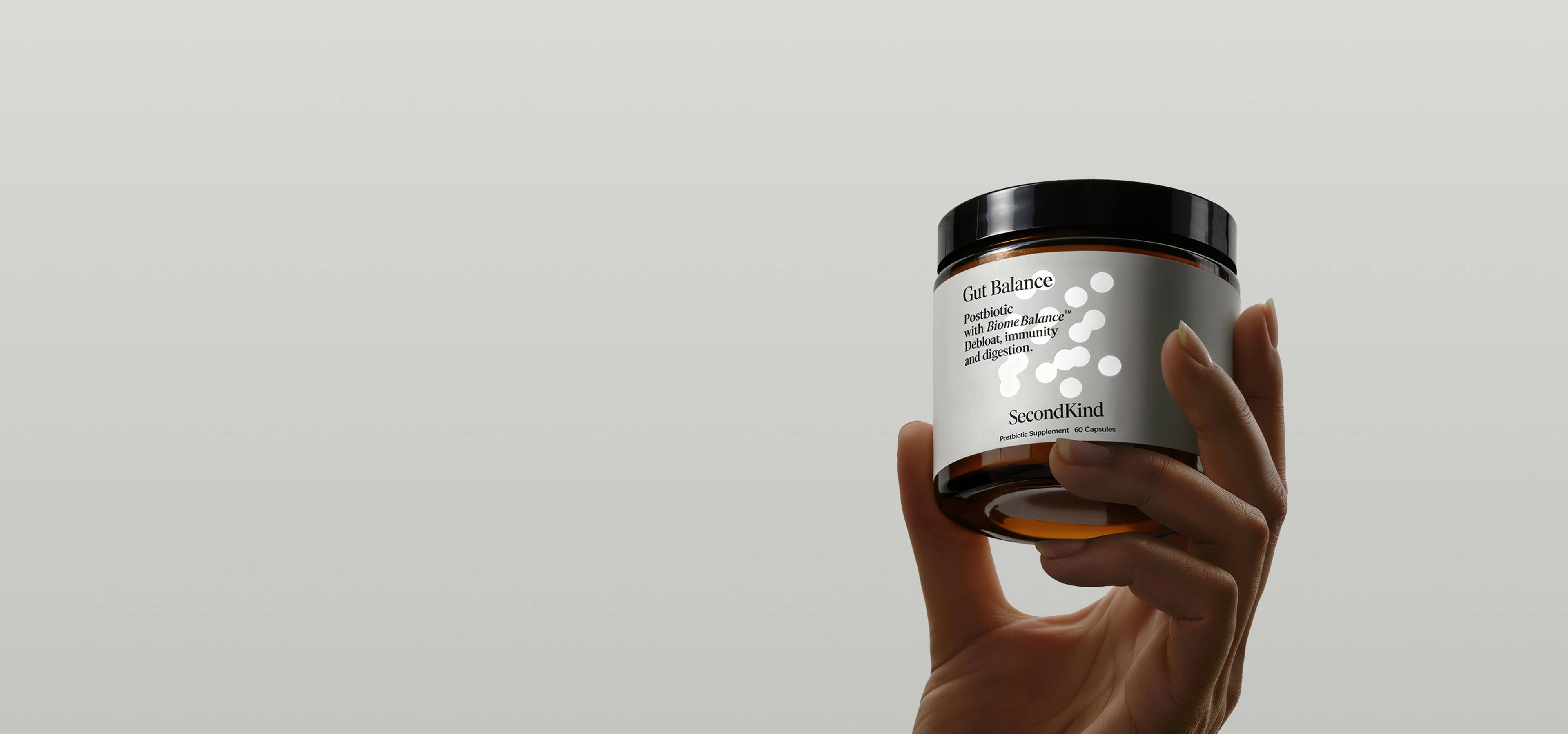 A hand holds up a brown jar labeled Gut Balance by SecondKind against a plain light gray background. The label features a minimalist white dot design and product details in black text.