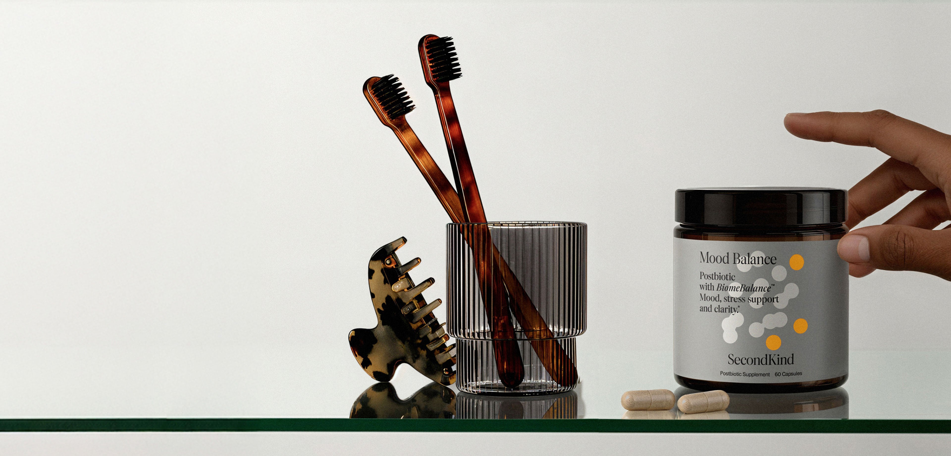 A hand reaches for a jar labeled Mood Balance beside a glass with two brown toothbrushes, a tortoiseshell hair clip, and two white capsules on a white surface.