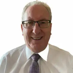 Smiling older man with short gray hair and glasses, wearing a white shirt and a patterned tie, against a plain white background.