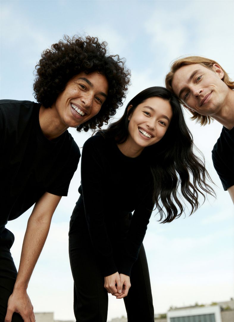 Three young adults, two men and one woman, wearing black shirts, smile and lean towards the camera outdoors against a bright, slightly cloudy sky.