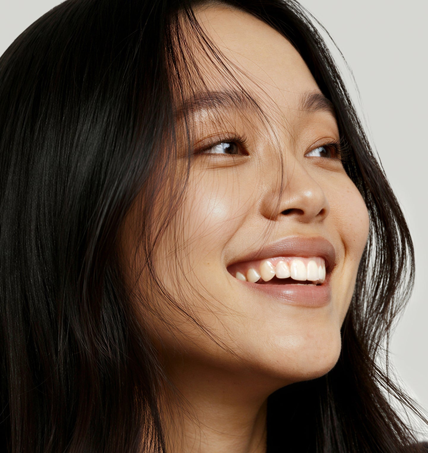 A young woman with long black hair smiles brightly, looking upward. She has clear skin and natural makeup. The background is plain and light-colored.