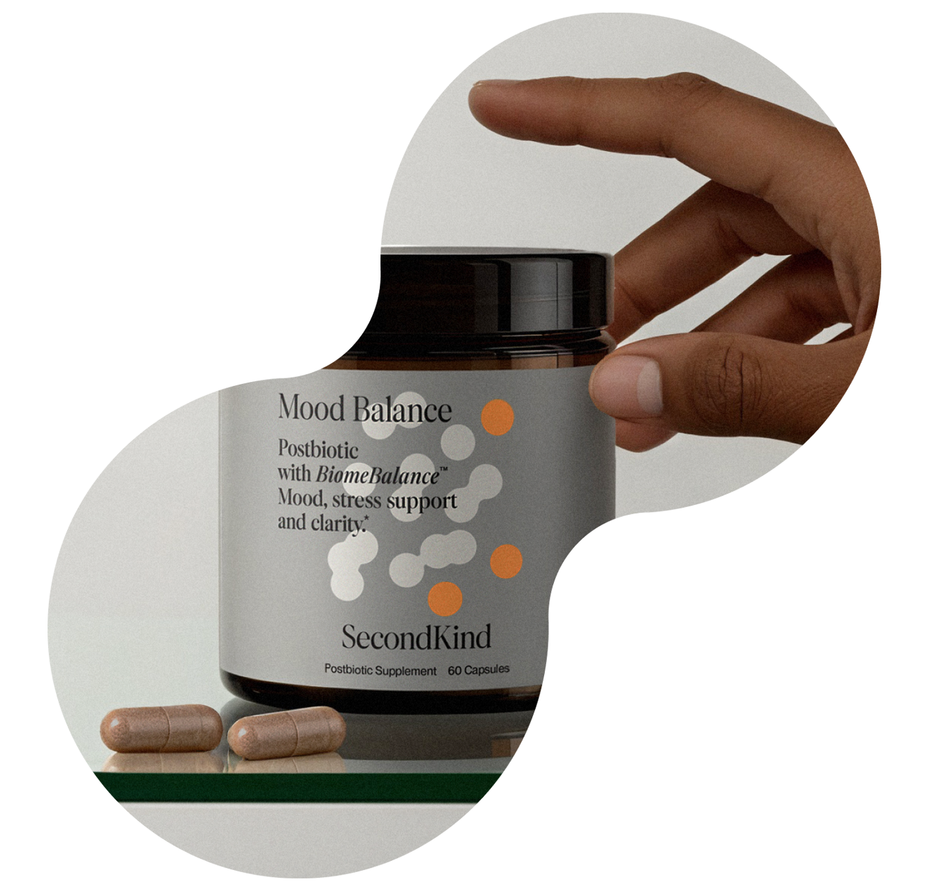 A hand holds a jar labeled Mood Balance by SecondKind, a postbiotic supplement for mood, stress support, and clarity. Two capsules are placed beside the jar on a white surface.