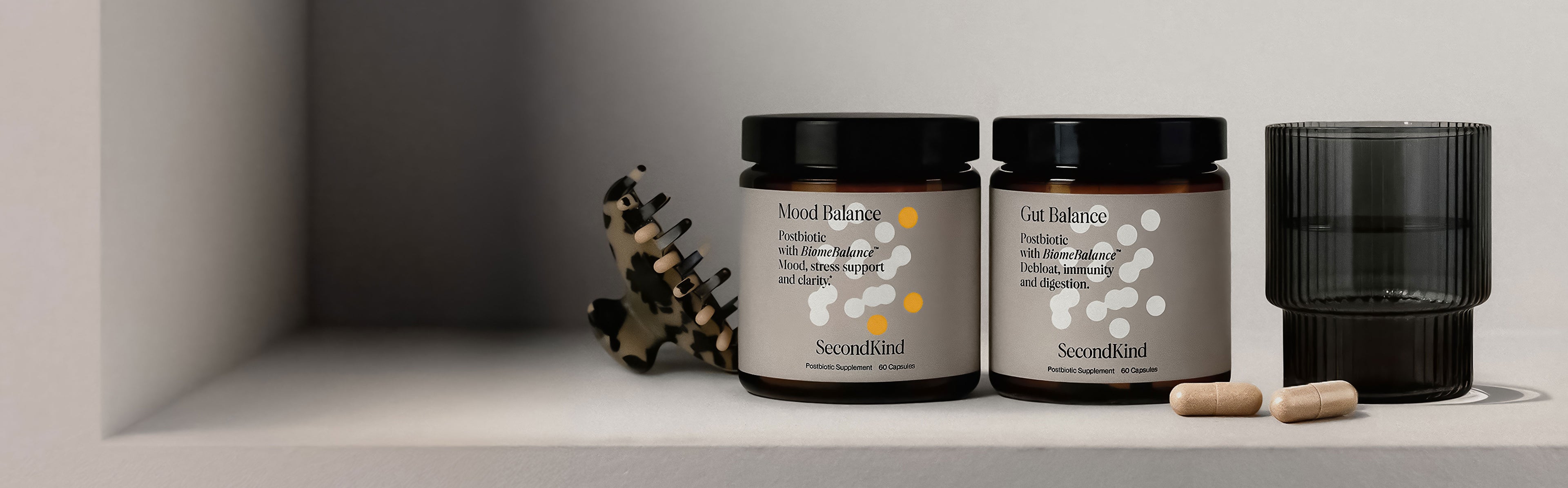 Two brown supplement jars labeled “Mood Balance” and “Gut Balance” from SecondKind sit on a shelf next to two beige capsules, a black glass cup, and a tortoiseshell hair clip.