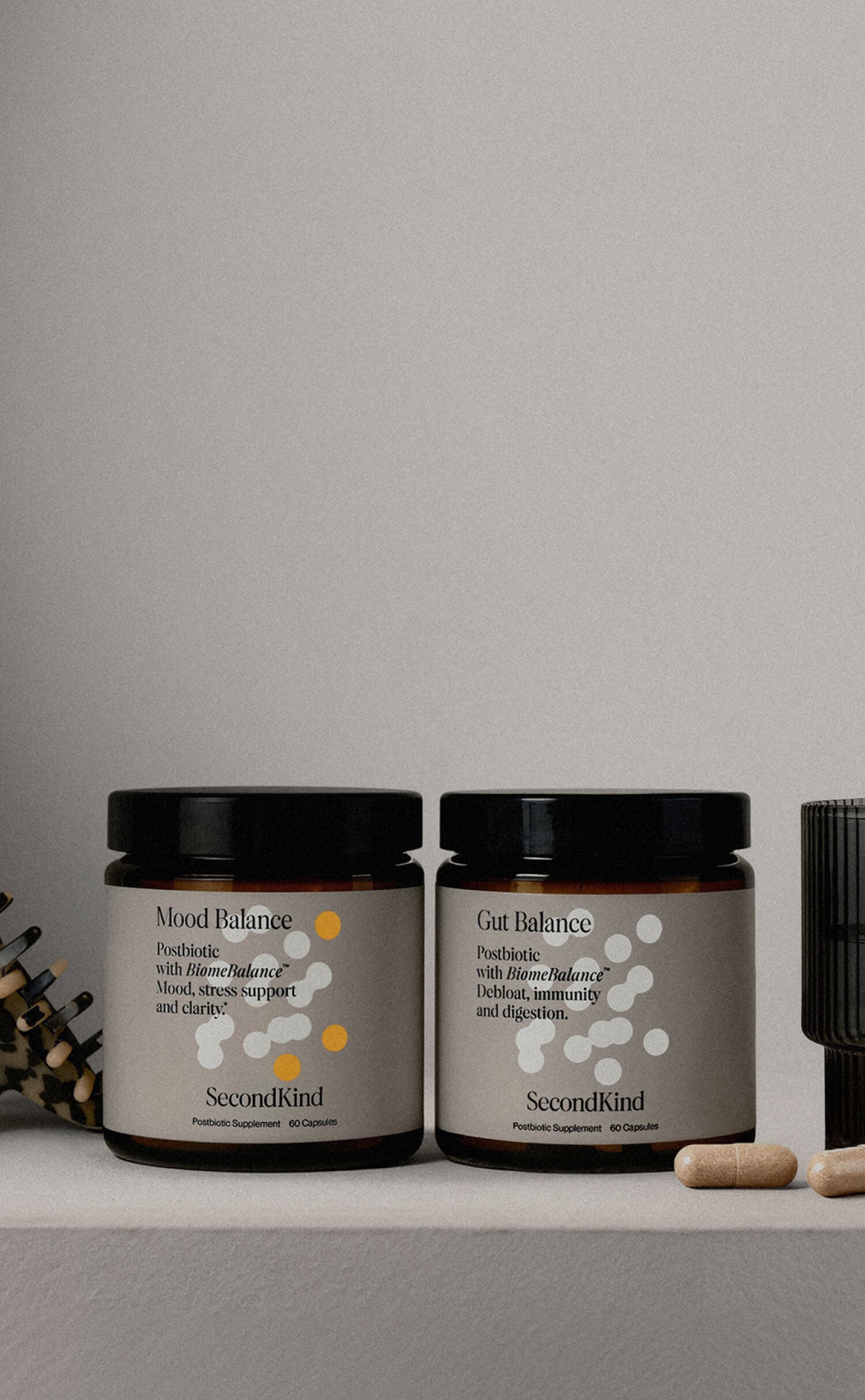 Two brown glass jars labeled Mood Balance and Gut Balance from Second Kind sit on a surface, with capsules, a notebook, and a black ribbed container nearby. The jars are dietary supplements.