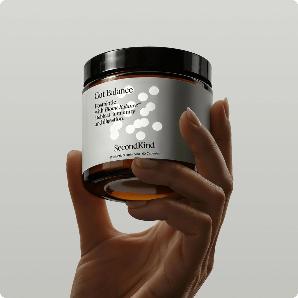 A hand holds a dark amber jar labeled Gut Balance, a postbiotic supplement supporting digestion and immunity, featuring white circles and minimalist design on a gray label.