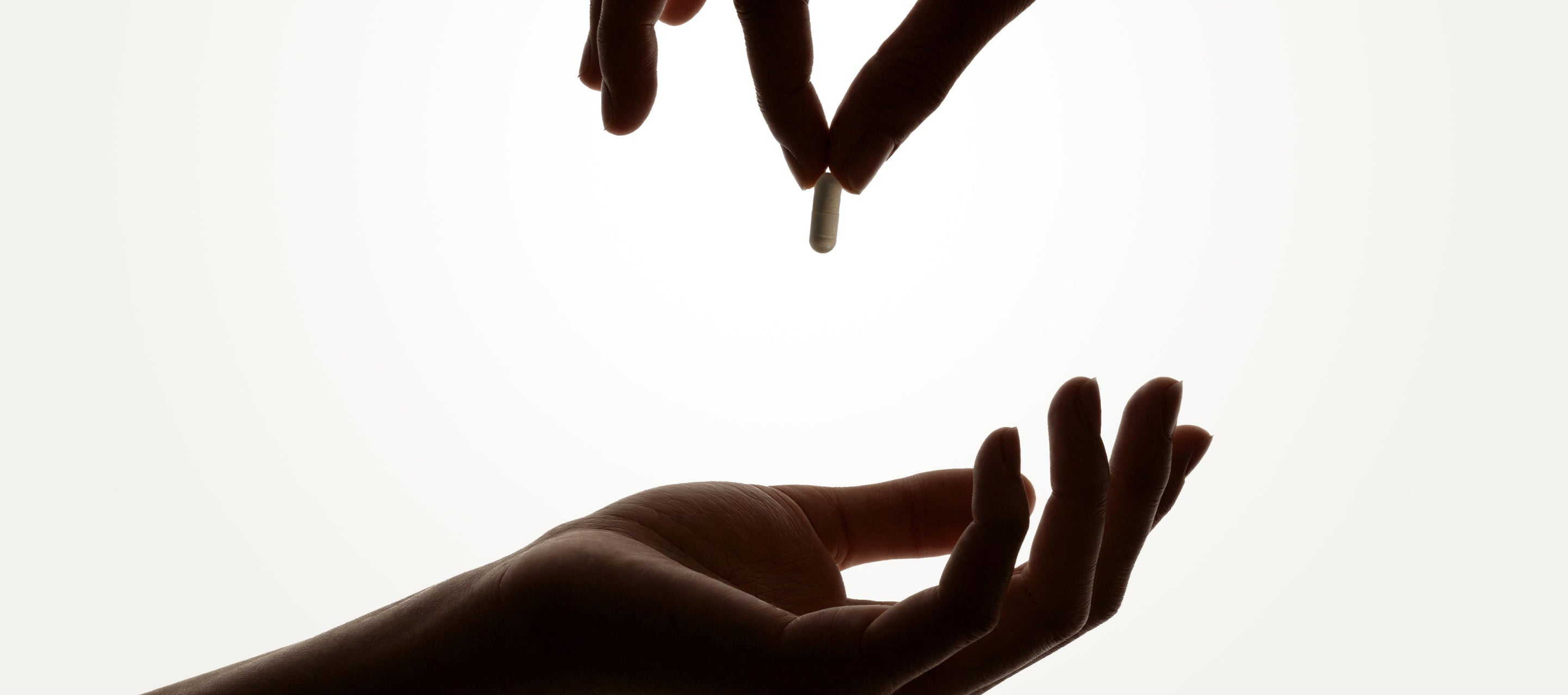Two hands silhouetted against a white background; one hand is holding a capsule-shaped pill and is about to drop it into the open palm of the other hand below.