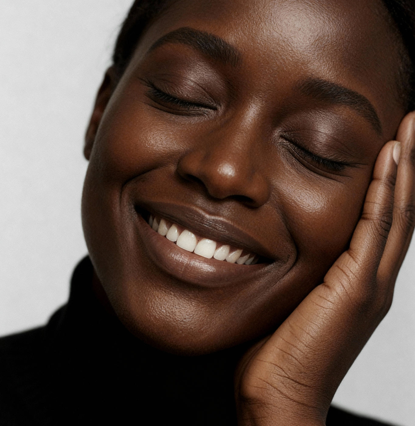 A close-up of a woman with dark skin smiling softly, eyes closed, and gently resting her face on her hand. She wears a black turtleneck and appears relaxed and content against a plain background.