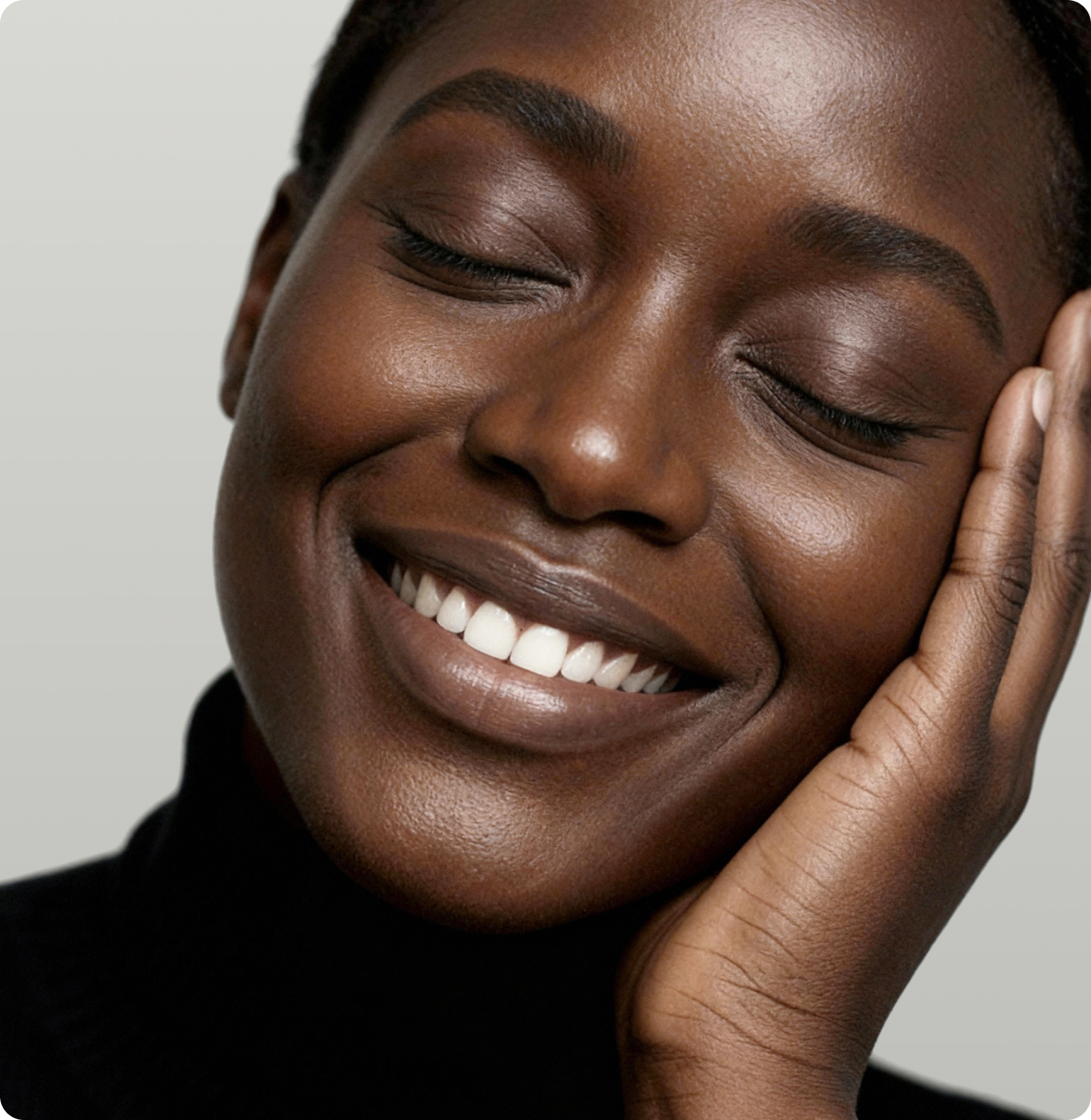 A person with dark skin smiles gently with their eyes closed, resting their face on their hand. They are wearing a black top against a light gray background.