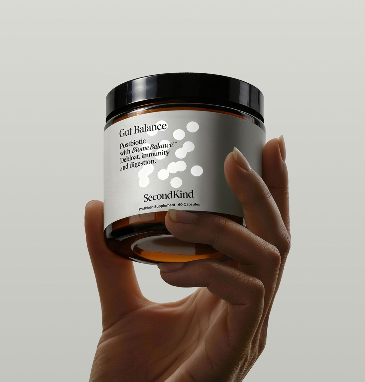 A hand holds a brown jar labeled Gut Balance with white dots on the label. The jar is a postbiotic supplement by SecondKind, described for digest, immunity, and digestion support. The background is plain and light-colored.