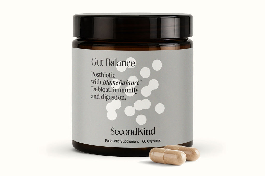 A brown jar labeled Gut and Mood Balance Bundle by SecondKind with white text describes a postbiotic supplement for digestion and gut–brain axis support, with two beige capsules in front of the jar.