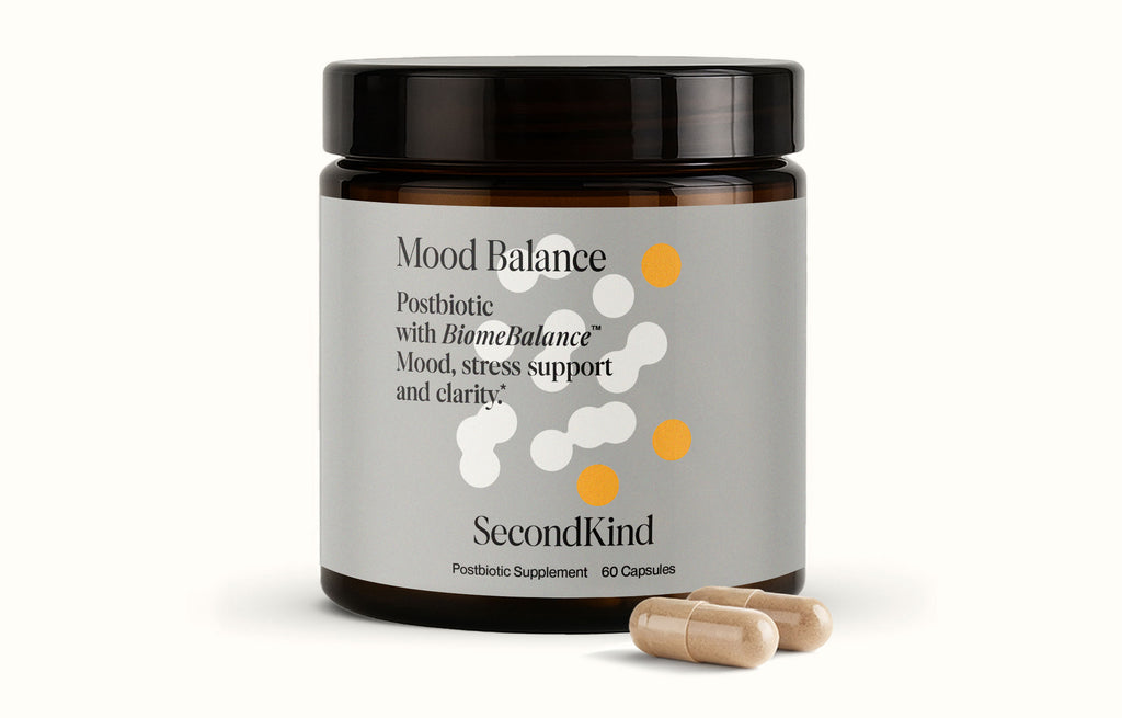 A brown jar labeled "Mood Balance" is shown with text for mood support, stress relief, and clarity. Two beige capsules are placed in front on a white background, highlighting this postbiotic supplement.