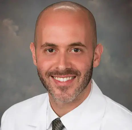 A smiling man with a bald head and neatly trimmed beard, wearing a white medical coat and a gray patterned tie, in front of a neutral gray background.
